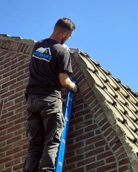 Vakman van Leermans in bedrijfsshirt klimt via een ladder langs de gevel naar een pannendak